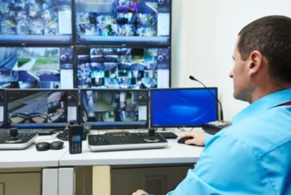 um homem de uniforme olhando câmeras de segurança e está com um celular na mão