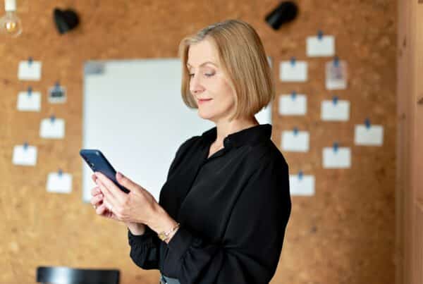 mulher em escrit[orio, está de pé segurando um cleular com as duas mão, olha para ele