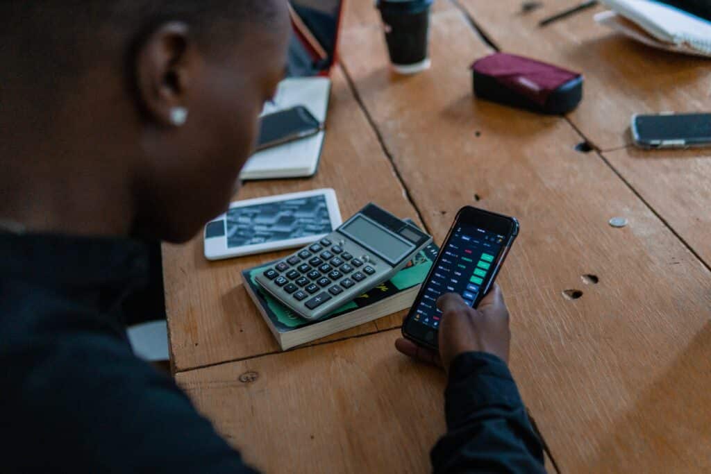 pessoa usa calculadora do celular, ao lado a uma calculadora em cima de um livro, sob a mesa
