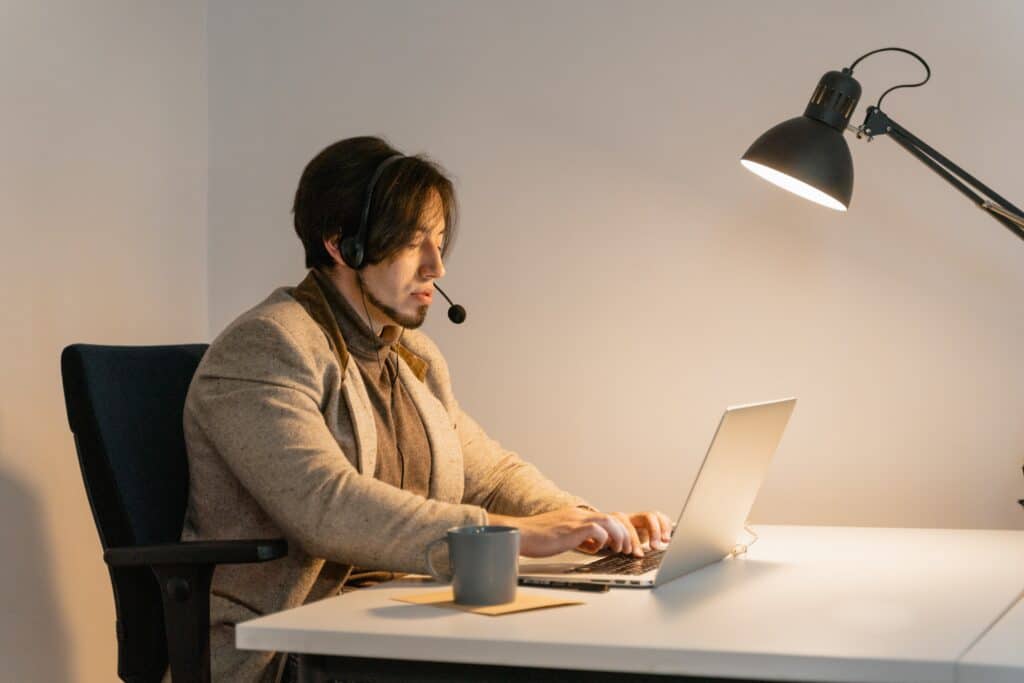 home sentado, digita em notebook com plataforma dispatcher, fala em headset, há uma xícara ao seu lado e um abajur a sua frente